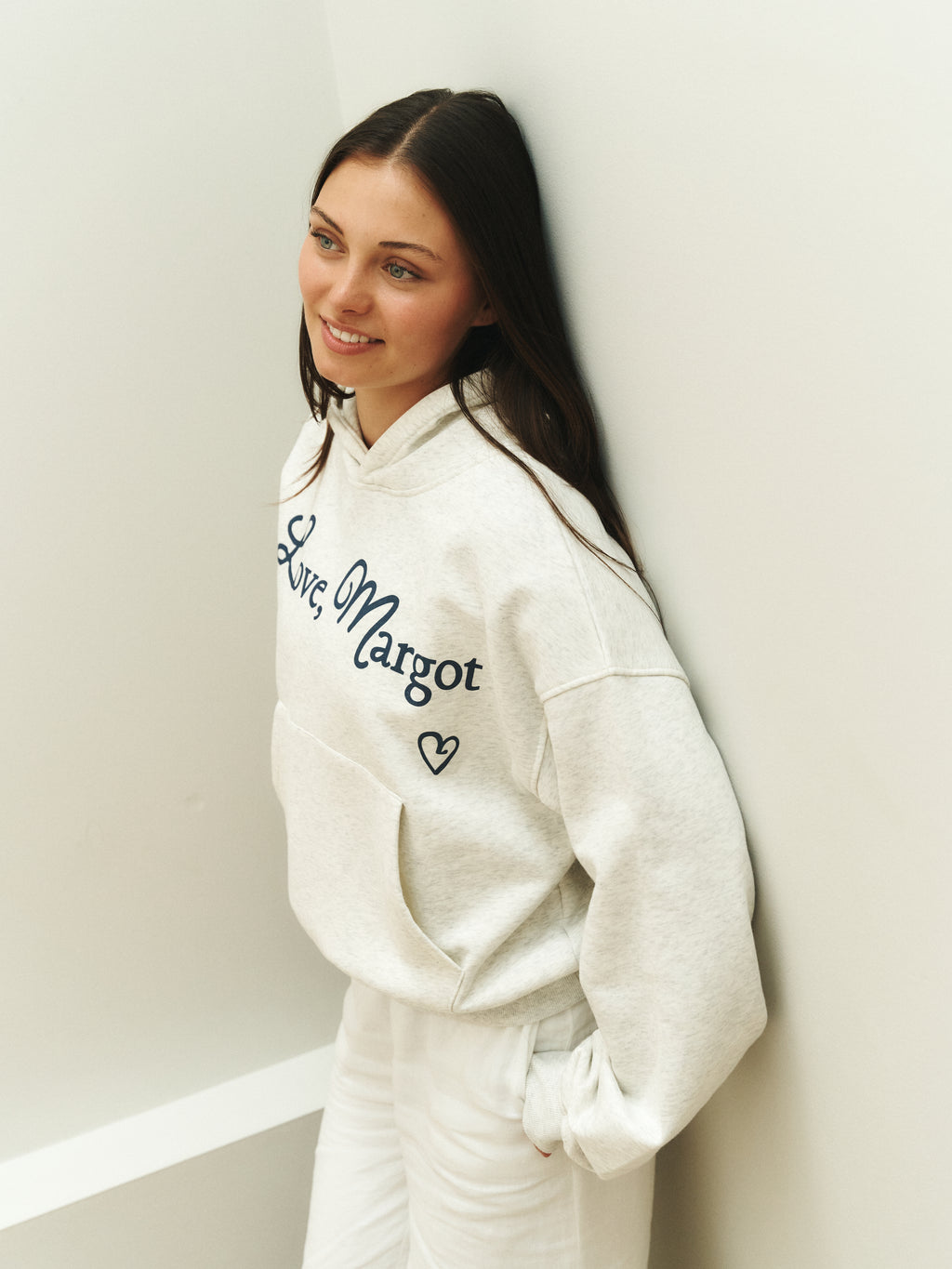Woman wearing a white hoodie with text and graphics, leaning against a light-colored wall.