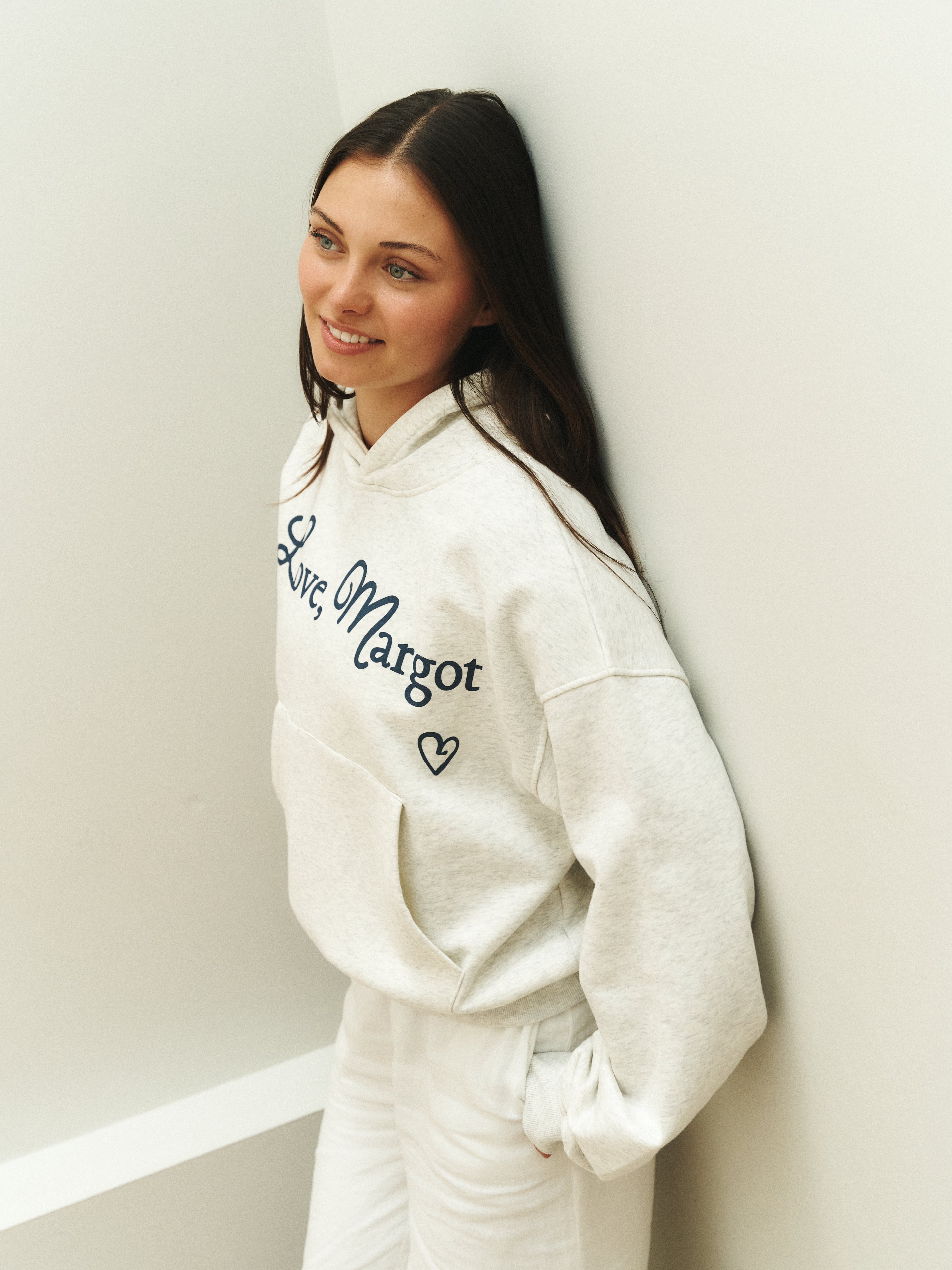 Woman wearing a white hoodie with text and graphics, leaning against a light-colored wall.